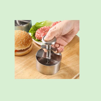 Presse à hamburger en inox sur une planche à côté d’un steak haché cru formé et d’un pain à burger.